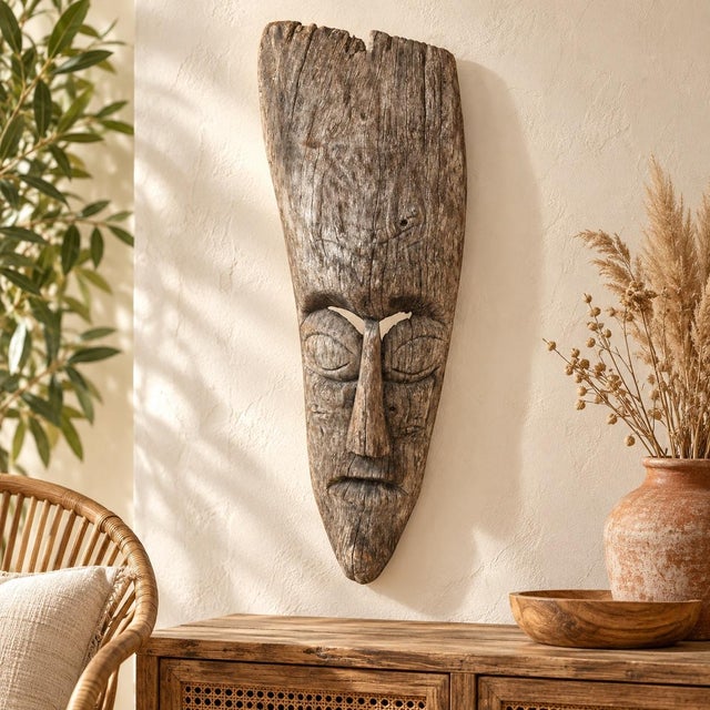 A striking carved wooden face mask with an elongated silhouette, deeply sculpted features, and a richly textured gray-...