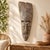 A striking carved wooden face mask with an elongated silhouette, deeply sculpted features, and a richly textured gray-...