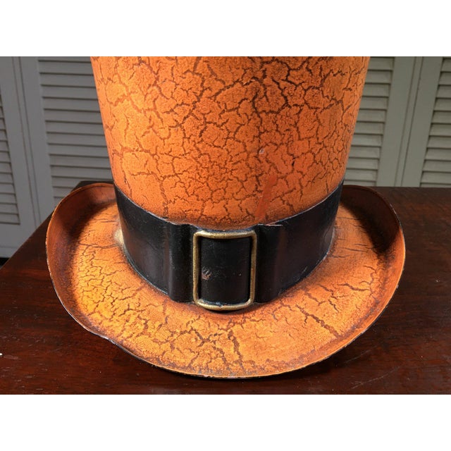 A decorative top hat with a bold copper-orange crackle finish and a black band accented by a brass buckle. The distressed,...