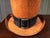 A decorative top hat with a bold copper-orange crackle finish and a black band accented by a brass buckle. The distressed,...