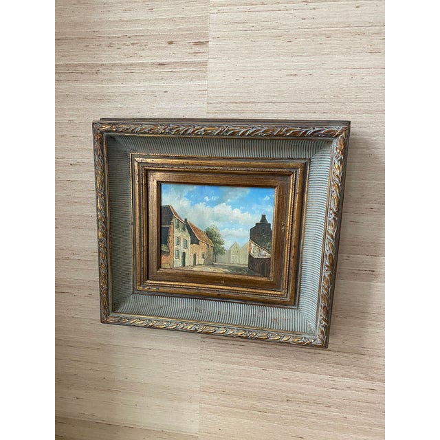Vintage oil painting with scene of hamlet or village in pretty blues and rust color. Substantial gold decorative frame.
