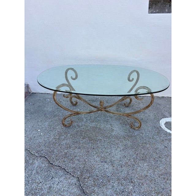 Vintage Hollywood Regency style coffee table featuring a gold-finished wrought iron base with scrollwork and an oval...