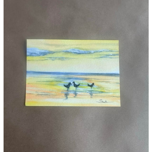 A serene, impressionistic watercolor capturing three shorebirds wading along a softly glowing shoreline. Rendered in a...