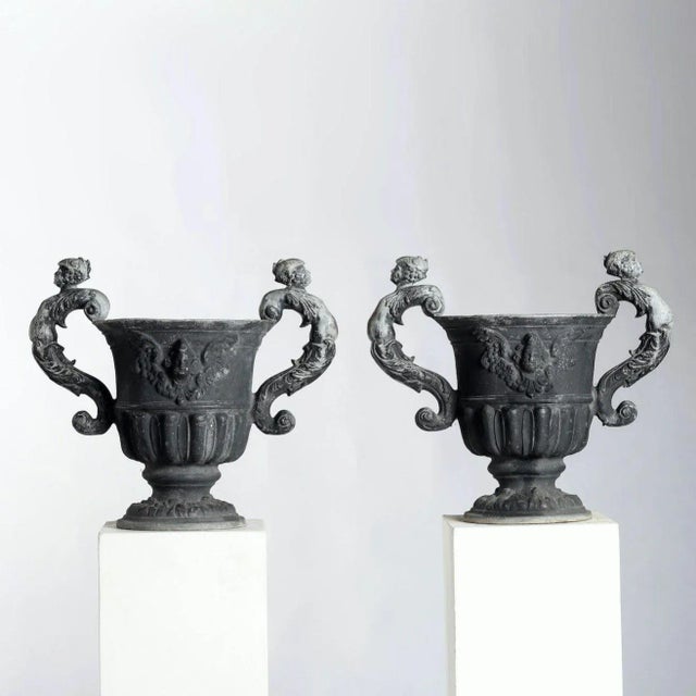Exceptional pair of English lead urns. Round, with a gadrooned waist, the sculpted lead depicts a winged cherub with...