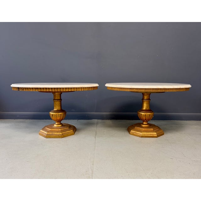 A refined pair of complimentary neoclassical-inspired side tables, each featuring a circular marble top with soft, natural...
