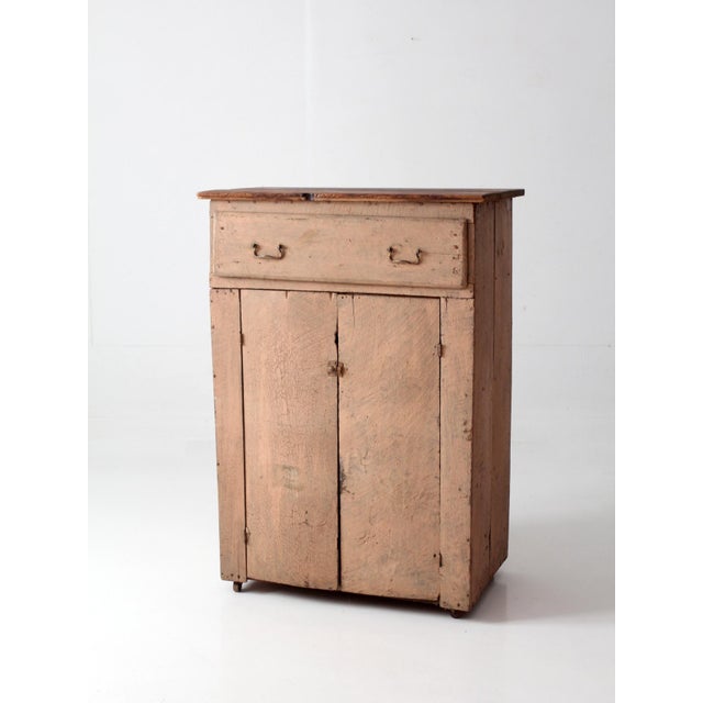 An antique painted wood cupboard featuring a timeworn faded peach finish, contrasting natural wood plank top, and...