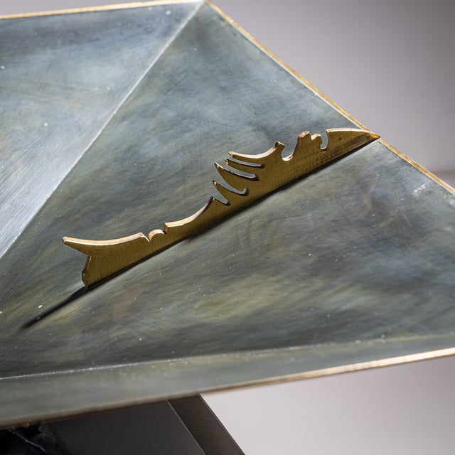 Square metal centerpiece raised on a pyramidal base, featuring a jagged decorative element along the rim, executed in a...