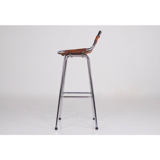 A bar stool with a slightly raised back, often attributed to charlotte perriand. Cognac leather seat riveted to a chromed...