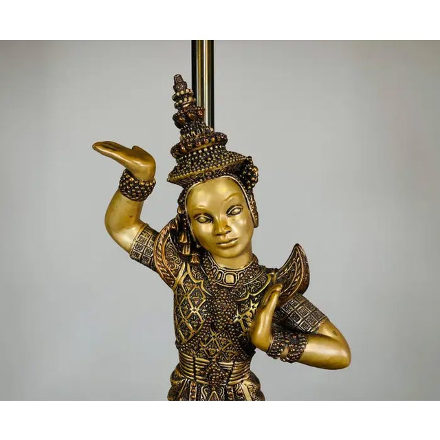 This beautiful lamp titled “Bangkok” features a golden statue of a Thai dancer as its base, holding up a dark, textured...