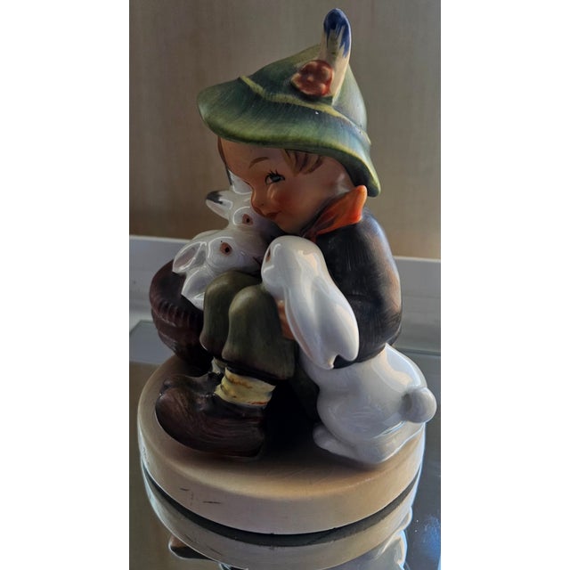 A charming vintage Goebel Hummel figurine titled “Playmates,” depicting a young boy gently holding two bunnies. Designed...