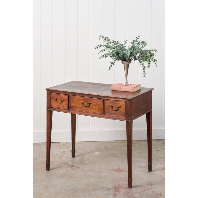 This early 19th-century oak three-drawer side table features three drawers adorned with brass swan-neck pulls. Ideal as a...