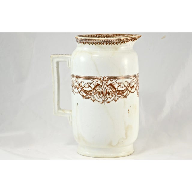 Antique brown English transferware pitcher encircled with an ornate scroll and Bacchus-like character pattern. The rim and...