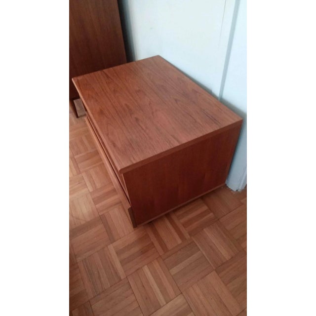 A low, wide teak nightstand in the Danish modern manner, with an open shelf above a full-width drawer. It rolls on hidden...