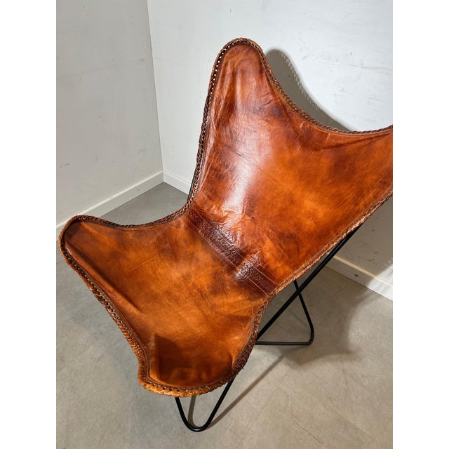 Classic butterfly chair featuring a rich cognac leather sling seat on a sleek black iron frame. A timeless mid-century...