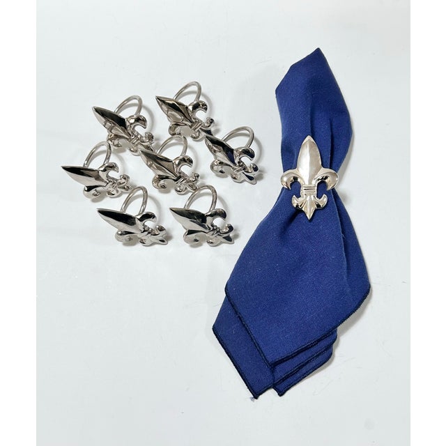 Set of eight vintage silver-tone metal napkin rings featuring sculptural fleur-de-lis designs with a polished, reflective...