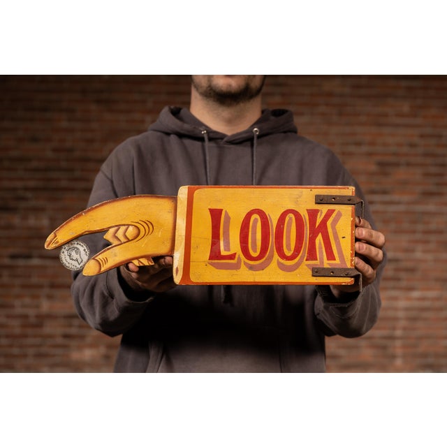 A vibrant hand-painted directional sign used to draw attention toward a nearby coin-operated machine or attraction. The...