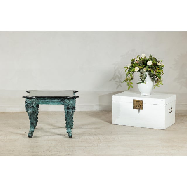 A striking bronze table with a rich verdigris patina and black marble top, its design marked by bold sculptural detail and...