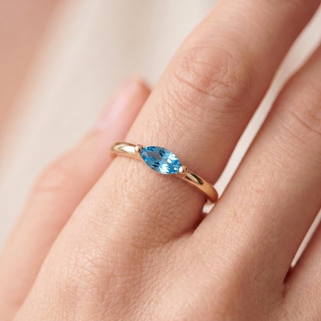 A cool, minimalist solitaire featuring a brilliant blue marquise-cut topaz set in an east-west orientation, held by a...