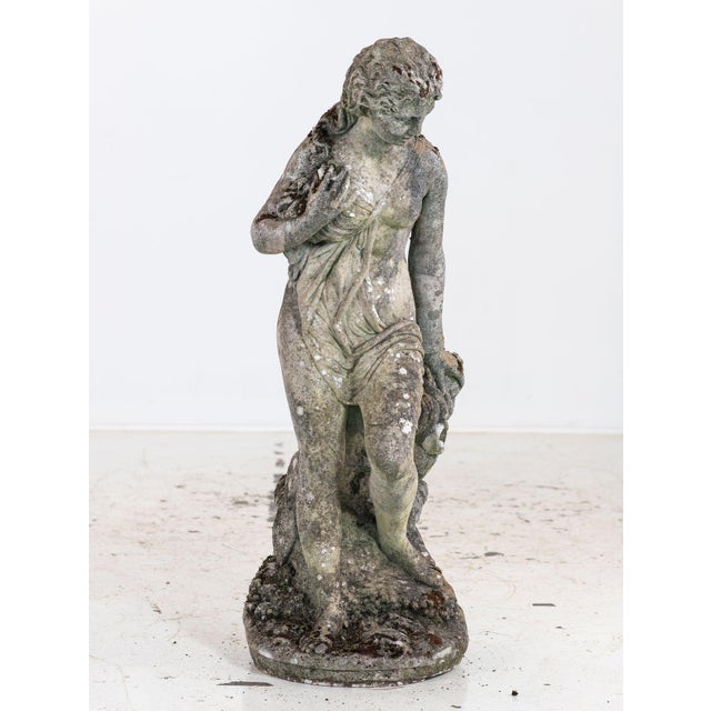This weathered composition stone statue depicts a maiden holding flowers or a draped garment. The patina of moss and age...