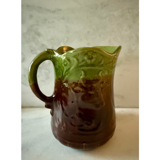 Vintage green and brown two-toned Majolica ceramic pitcher featuring embossed floral, leaf, and cow motifs.