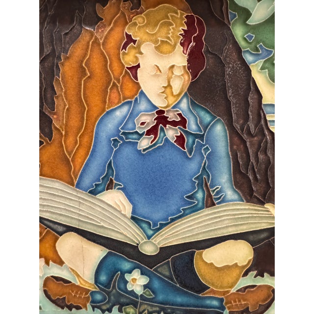 Decorative vintage glazed ceramic art tile depicting a seated boy reading an oversized open book beneath a tree, rendered...