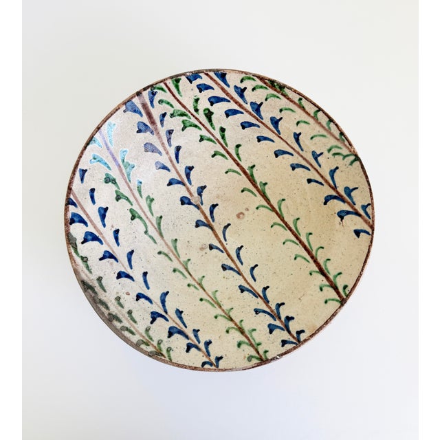 An antique 19th century earthenware bowl with hand-painted blue and green botanical brushwork. Crafted from a warm...