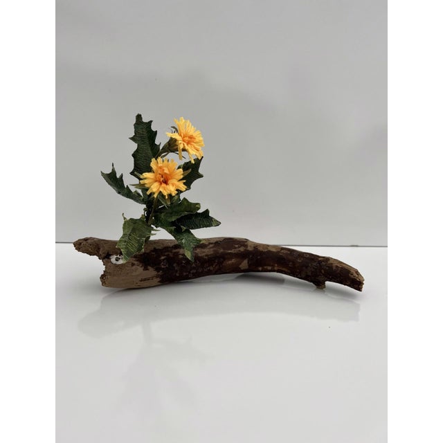 Kindling features a piece of driftwood that serves as a natural base for vibrant yellow flowers, juxtaposed against the...