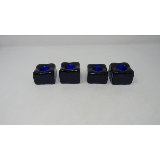 A set of four cobalt blue glass candlestick holders. The holders are small squares of glass with a knobby underside. They...