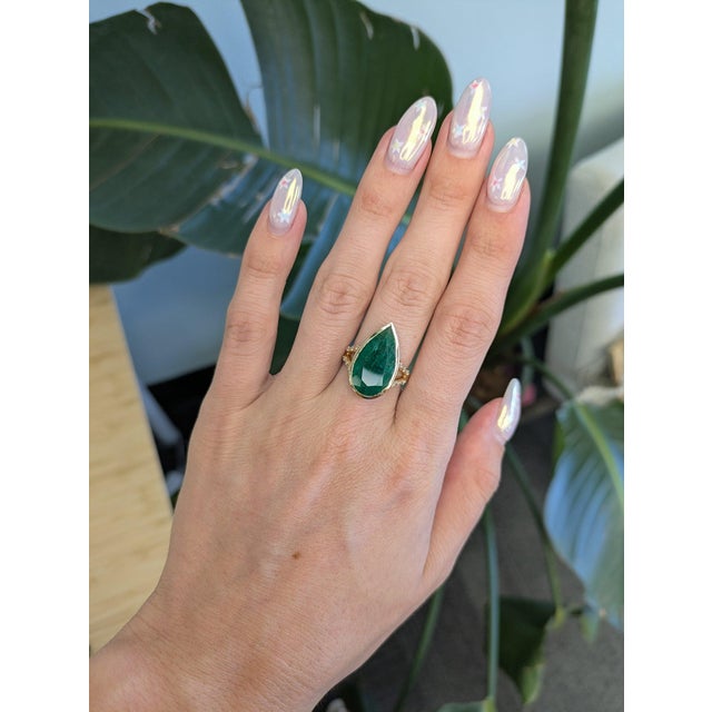 A lush Zambian emerald glows at the heart of this pear-shaped ring, its vivid green framed by a delicate trail of...