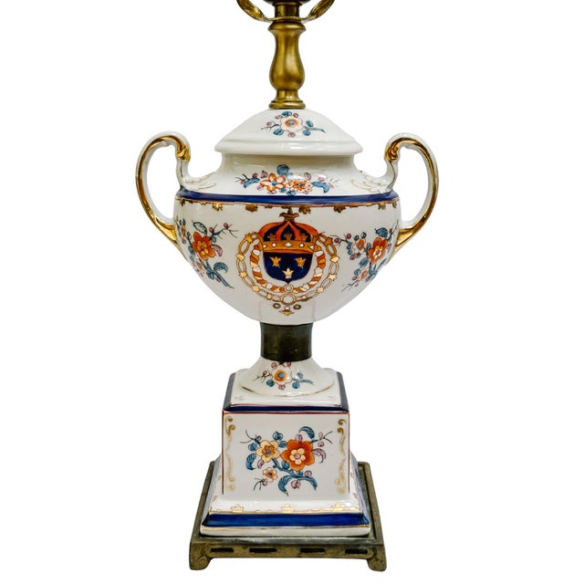 A sophisticated vintage porcelain table lamp featuring a classic handled urn-shaped body mounted on a footed brass base....
