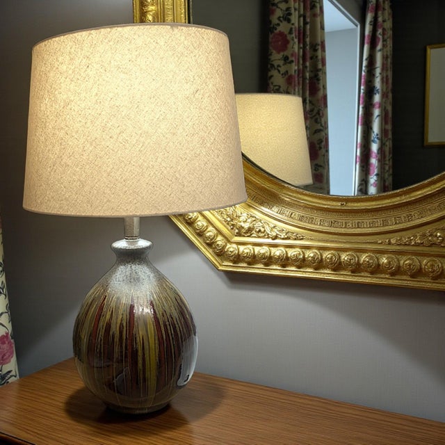 An artisanal mid-century drip glaze mannered ceramic lamp is displayed in tones of olive green, mustard yellow, and...