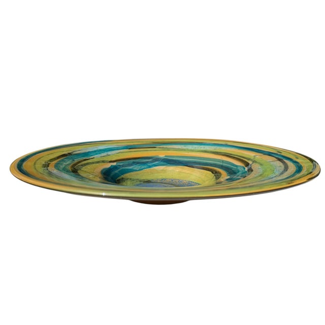 Large handcrafted art glass charger bowl featuring swirling bands of cobalt blue, teal, chartreuse, amber, and black. The...