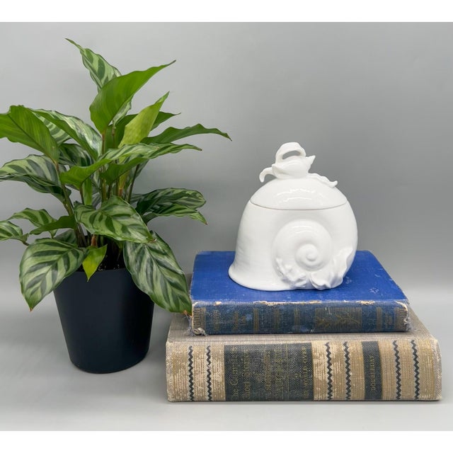 Whimsical white porcelain lidded jar in the shape of a spiral snail shell, topped with sculpted seaweed and a tiny conch....