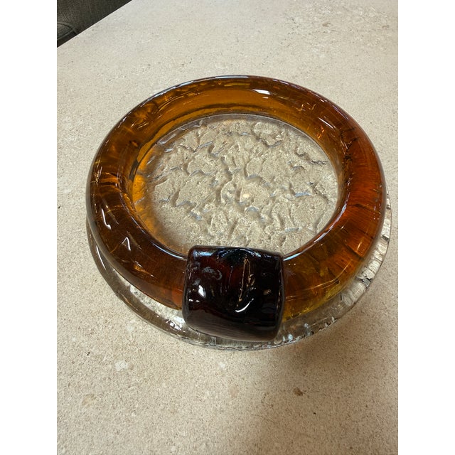 Striking vintage ashtray crafted from thick amber-toned glass with a clear, textured base. The piece features a bold,...