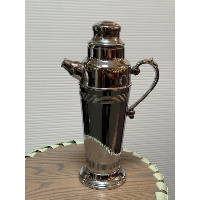 Design Plus Gallery presents a Vintage Art Déco Style Silver-Toned Cocktail Shaker. The shaker has a flared foot,...