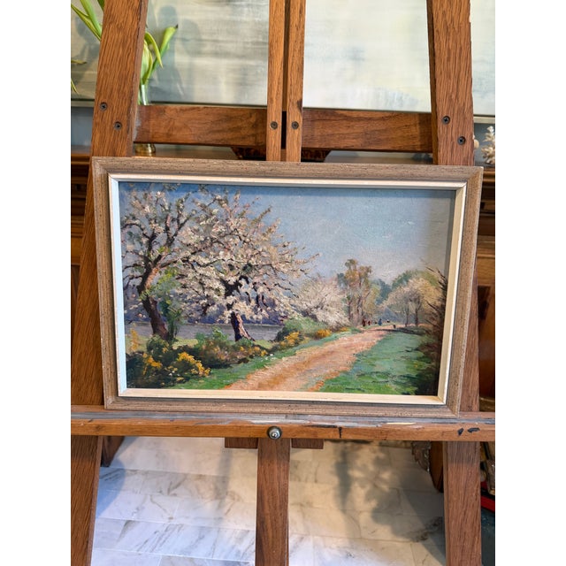 A luminous spring landscape — apple or cherry trees in full white blossom arch over a sunlit path running along a river,...