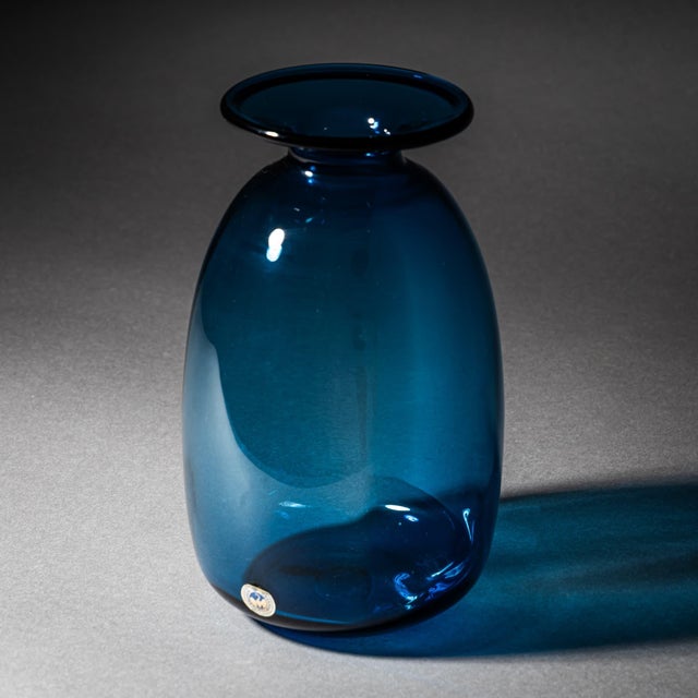 Small glass vase in an attractive petrol blue tone, featuring an elongated, rounded body, a narrow neck, and a wide lip....