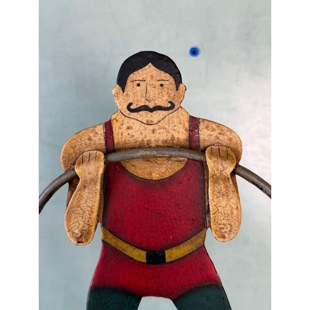 A charming vintage folk art sculpture depicting a classic carnival strongman performing a balancing act. The figure,...