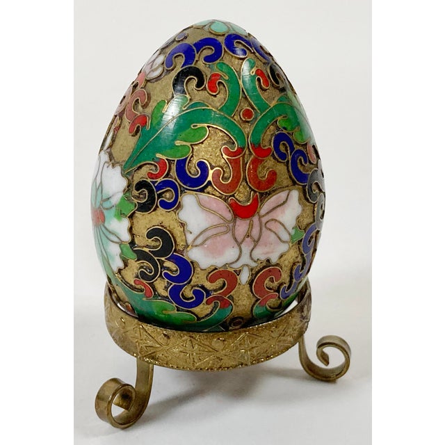Brass scrolled foot stand presents decorated enamel Cloissone egg. Egg has vibrant colors of navy, red, green, white, and...