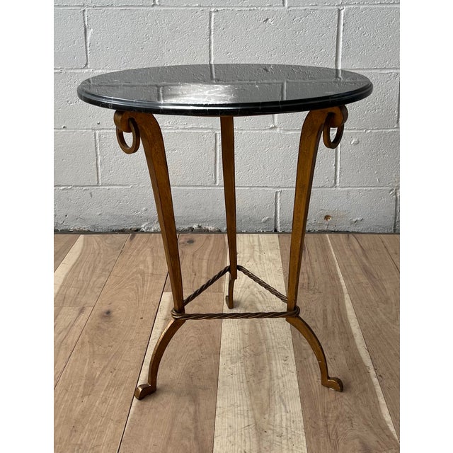 A refined Art Deco–style round side table featuring a gilt iron base and a sleek black marble top. The circular stone...