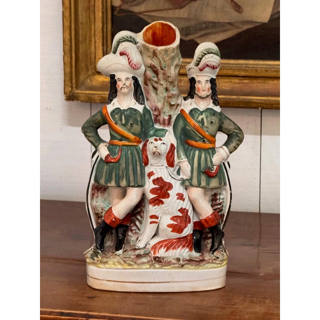 Two figures in green tunics and plumed hats stand either side of a tree trunk spill, with a red and white spaniel seated...
