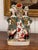 Two figures in green tunics and plumed hats stand either side of a tree trunk spill, with a red and white spaniel seated...