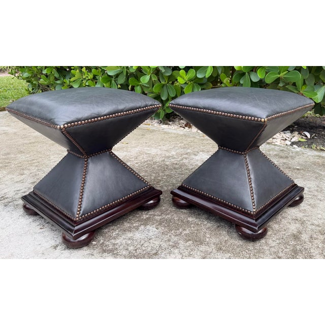 Vintage Baker leather footstool pair featuring a distinctive hourglass silhouette, dark gray leather upholstery, and...