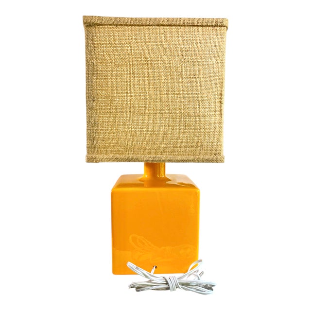 A vibrant orange ceramic table lamp with cube shaped body and square burlap shade.