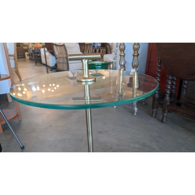 A petite spot table featuring a white marble base, brass stem and round glass top. The stem terminates in a small T shaped...