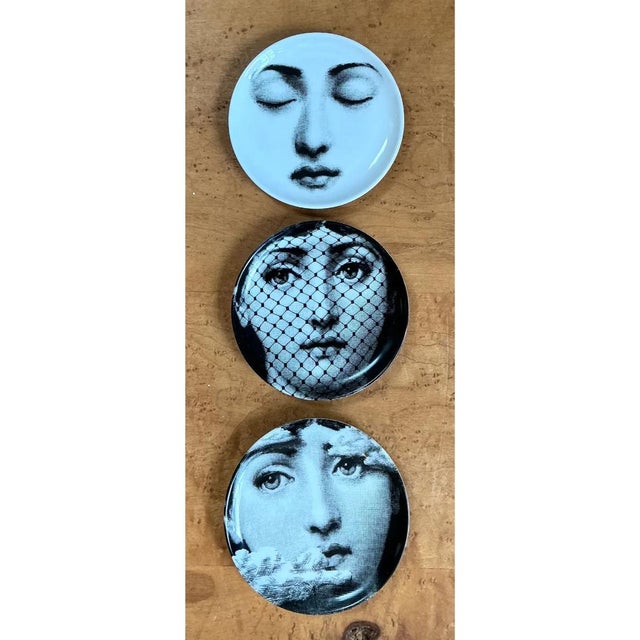 This set of three ceramic plates in the Fornasetti Tema e Variazioni style presents the designer's iconic recurring face...