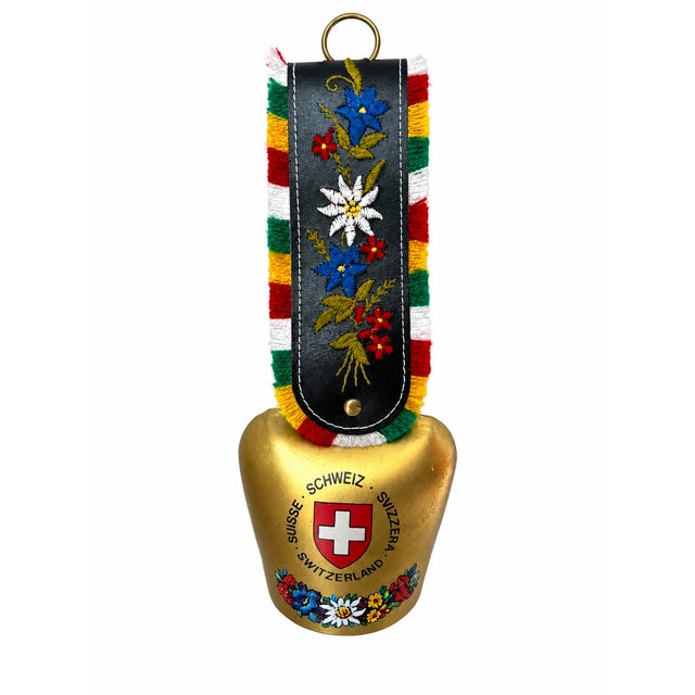 A charming Swiss-themed cowbell featuring a gold-tone metal body and a black strap embroidered with colorful Alpine...