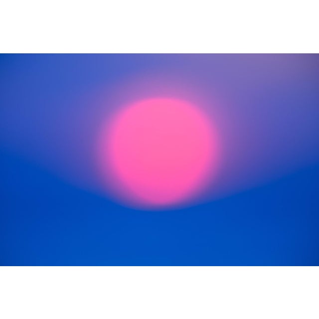 The artwork presents a soft, ethereal sunset, where a large, pink sun hovers against a gradient of blue, creating a...