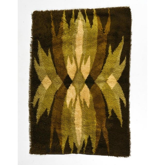 A hand-woven Scandinavian wool Rya tapestry features a symmetrical geometric pattern in earthy tones of green, brown,...