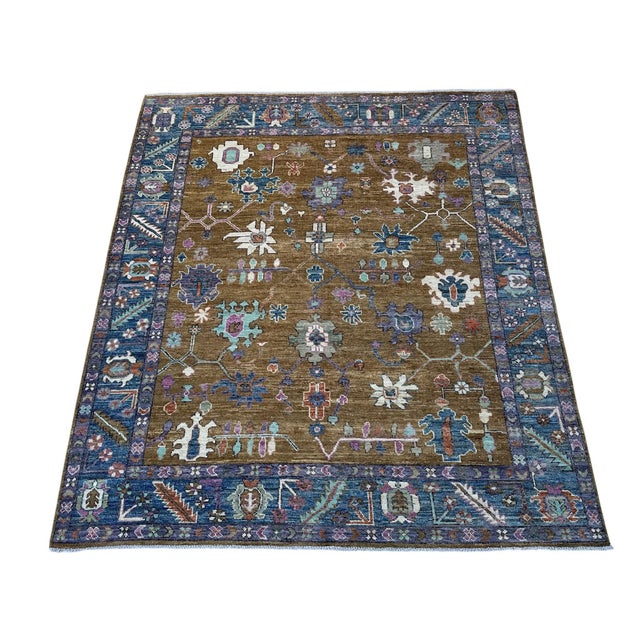 An olive-brown field and deep blue floral border distinguish this handmade Oushak wool rug. Scattered blossoms and angular...